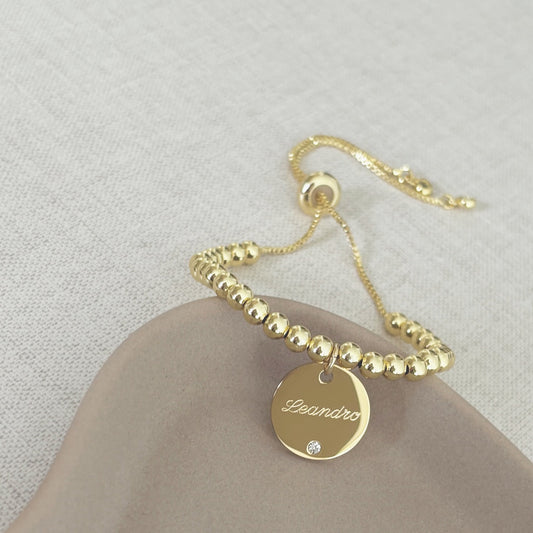 Pulsera formada por una cadena de color plateado con un dije de color plateado que tiene un nombre grabado en su superficie.