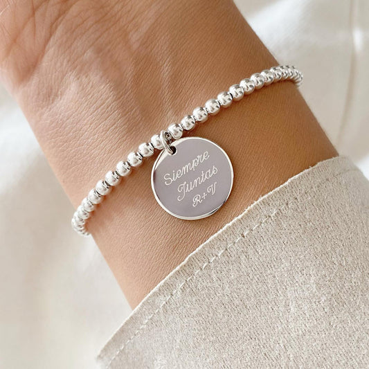 Pulsera formada por una cadena de color plateado con un dije de color plateado que tiene una frase grabada en su superficie.