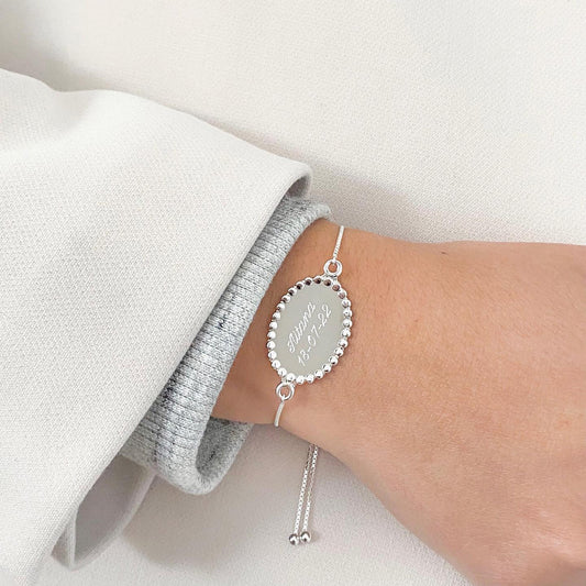 Pulsera formada por una cadena de color plateado con un dije de color plateado que tiene un nombre en su superficie.