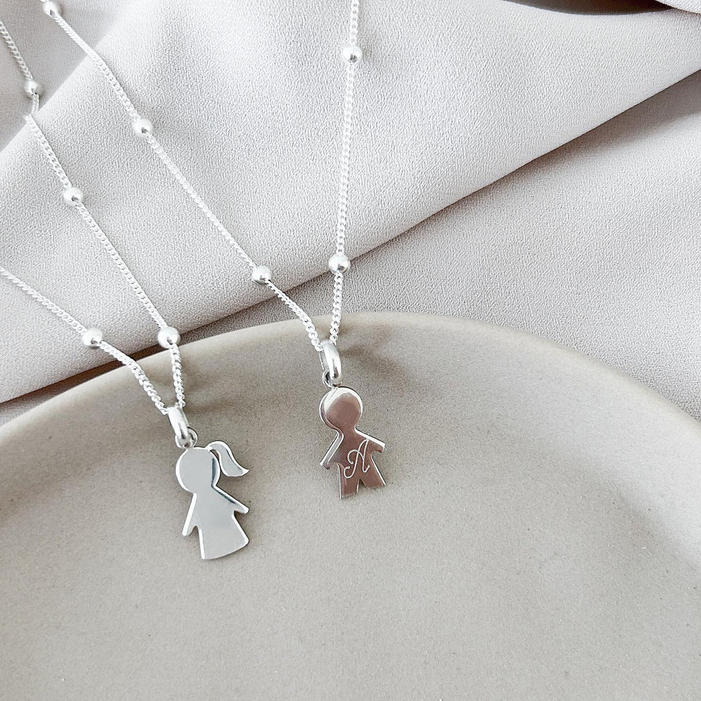 Dos collares, cada uno conformado por una cadena de color plateado. Uno de los collares tiene un dije de con la forma de la silueta de un niño de color plateado con una inicial grabada en su superficie. El otro collar tiene un dije con la forma de la silueta de una niña de color plateado.