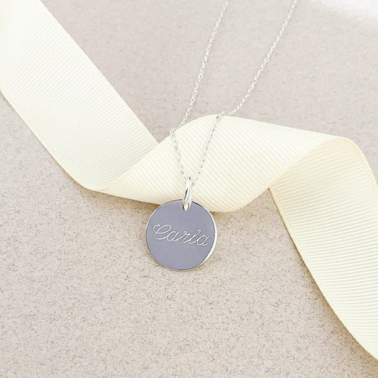 Cadena de color plateado con un dije de color plateado que tiene un nombre grabado en su superficie.