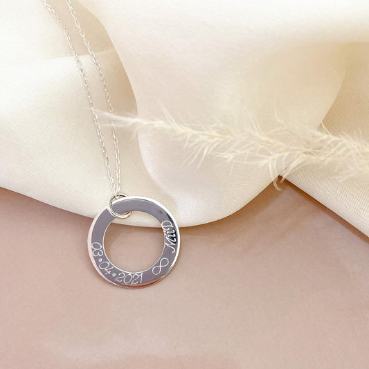 Pulsera formada por una cadena de color plateado con un dije redondo de color plateado que tiene una fecha y un nombre grabados en su superficie.