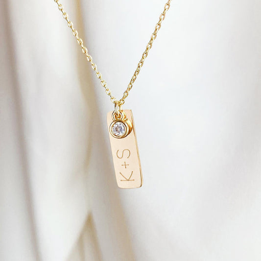 Collar formado por una cadena de color dorado con un dije con un cristal y uno de color dorado que tiene unas iniciales grabadas en su superficie. 