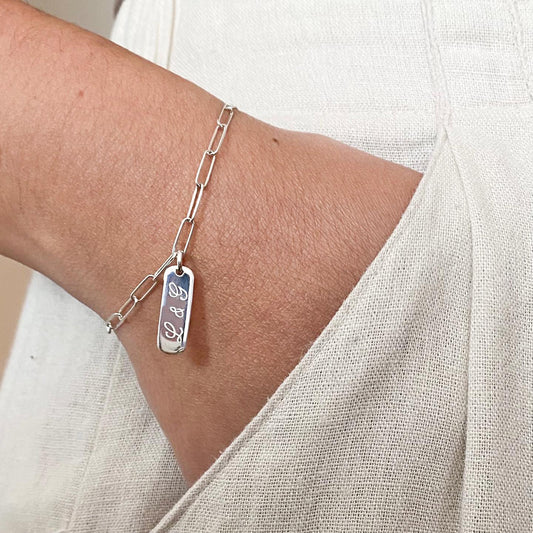 Pulsera formada por una cadena de color plateado con un dije de color plateado que tiene unas iniciales grabadas en su superficie.