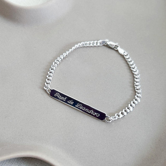 Pulsera formada por una cadena de color plateado con un dije de color plateado rectangular que tiene una frase grabada en su superficie.