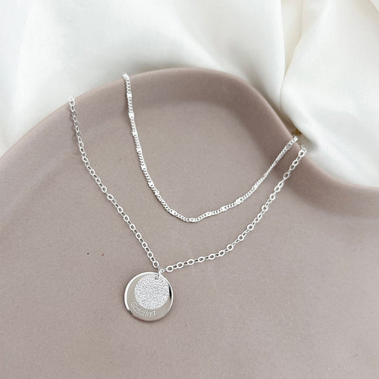 Collar formado por una doble cadena de color plateado con un dije de color plateado que tiene un nombre grabada en su superficie.